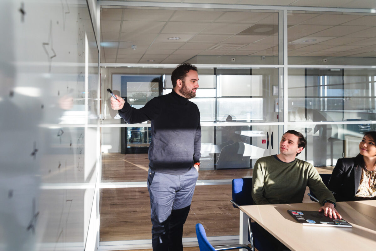 A person in front of a whiteboard pointing to it and two people in the same meeting room listening to him.