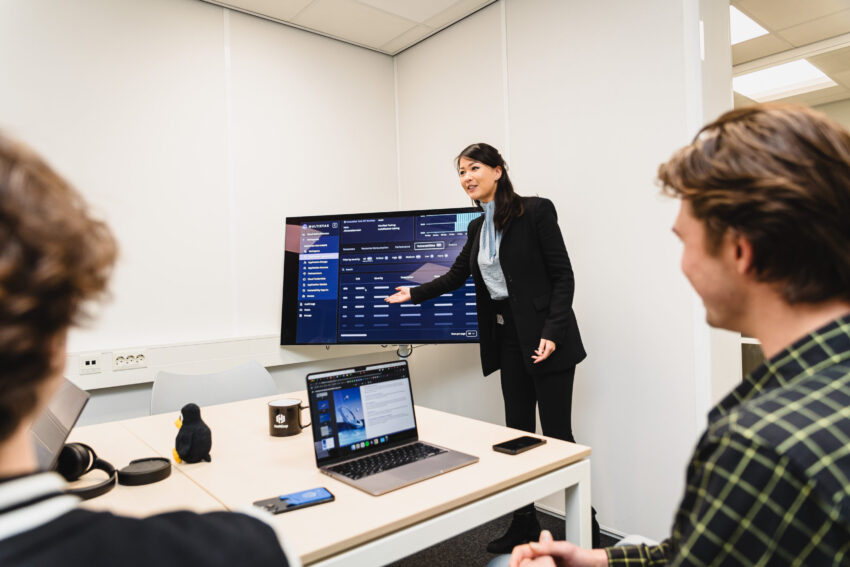 Three people in a meeting room, one standing in front of a screen.