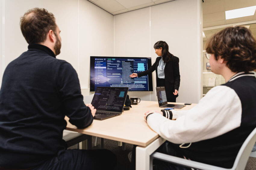 Three people in a meeting room. One is standing in front of a screen giving a presentation.