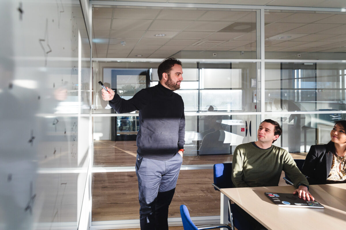 Three people in a meeting room. One person in front of a whiteboard.