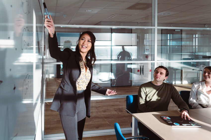 Three people in a meeting room. One person standing in front of a whiteboard pointing towards it.