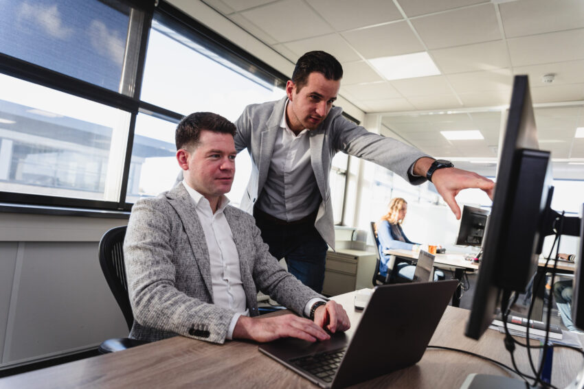 Two Sue people next to each other. One is typing on a laptop, the other one points to the screen. In the background is a third person behind a computer.