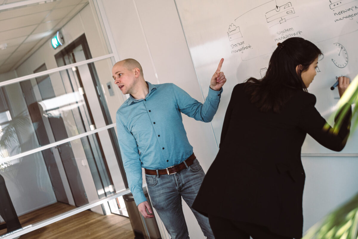 Two people in front of a whiteboard. One person pointing.