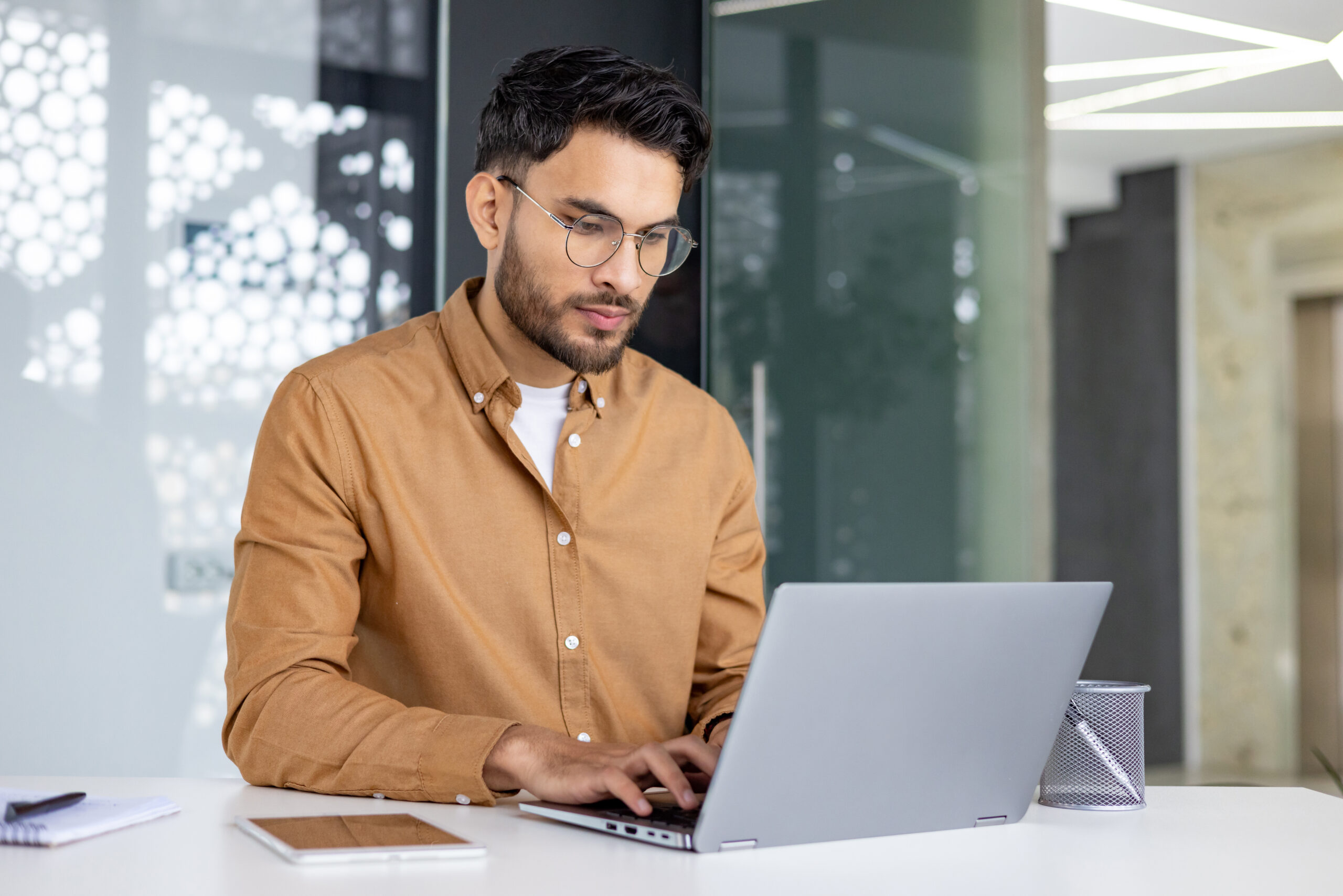 Young man in glasses working on a laptop in a stylish office setting. He appears concentrated and engaged in his tasks.